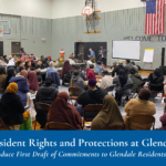 Community meeting in a gymnasium with people seated facing speakers and a presentation screen.