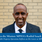 A smiling man in a suit stands in front of a tan brick wall.