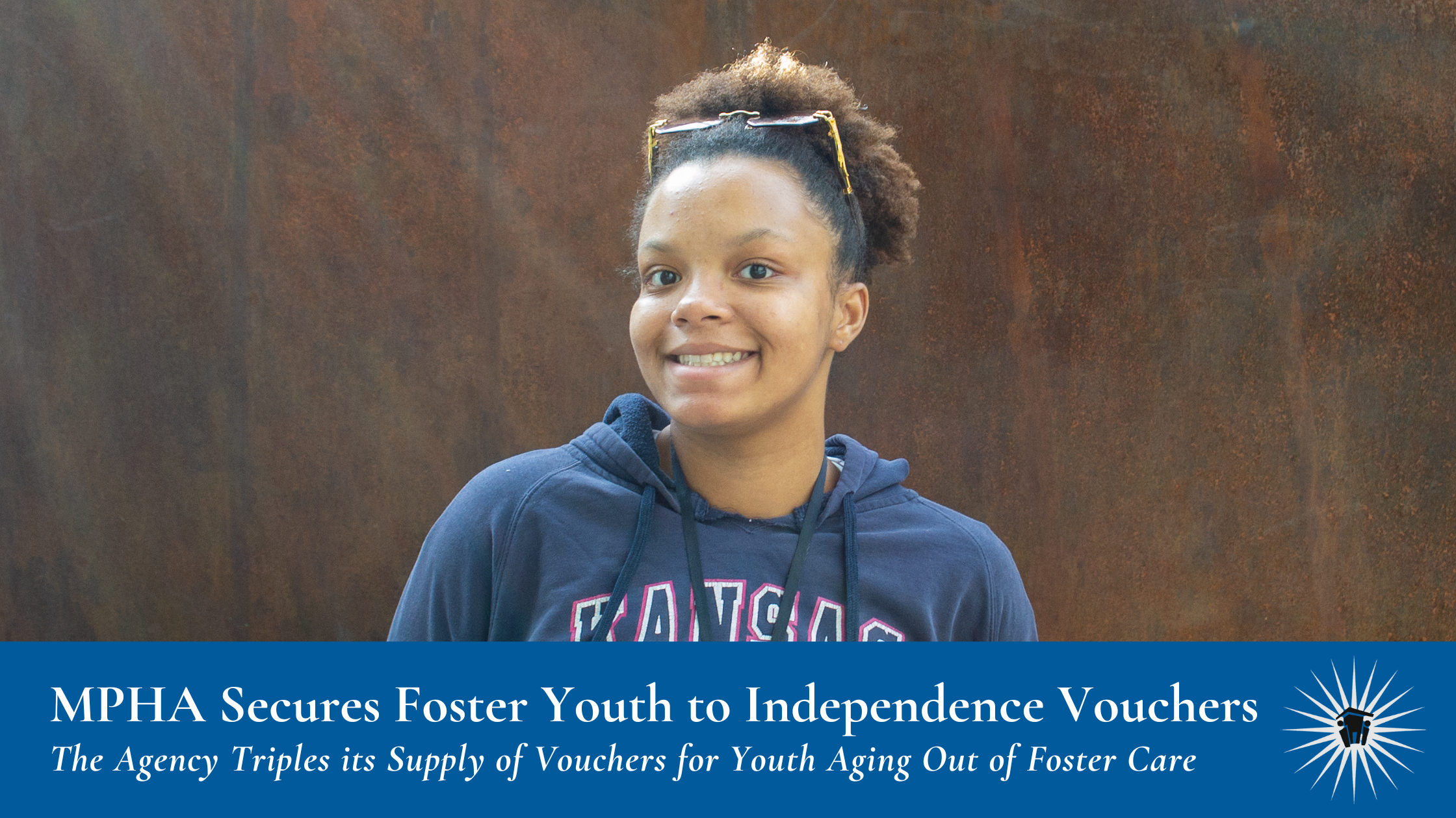 A young woman in a navy hoodie stands in front of a brown wall, smiling.