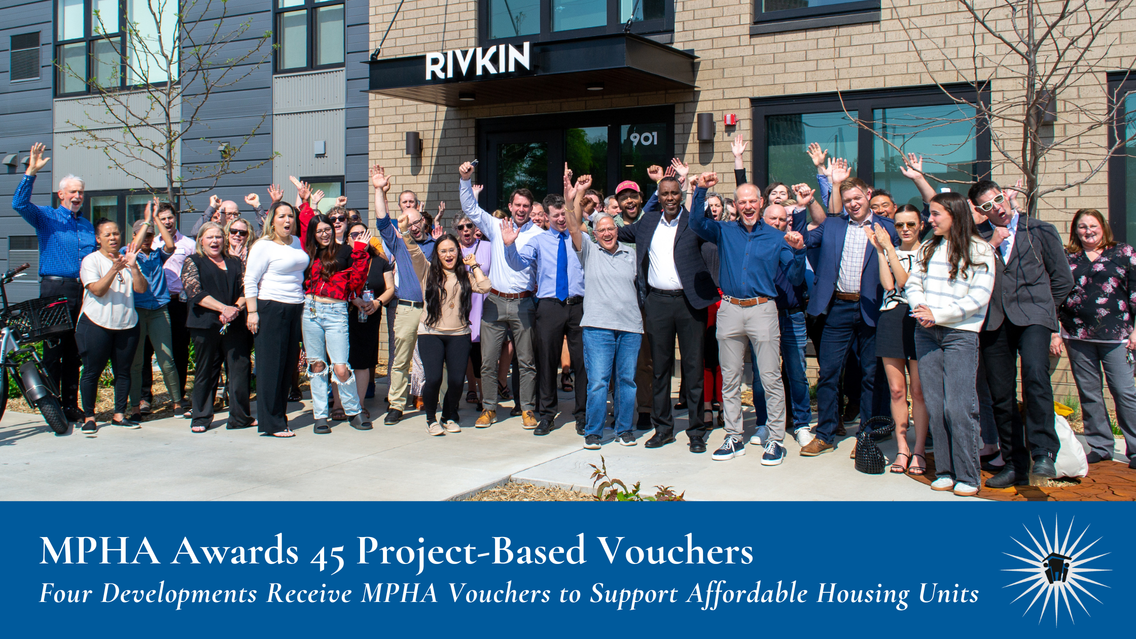 A large group of people stand and celebrate in front of a modern apartment building with a sign reading "Rivkin.