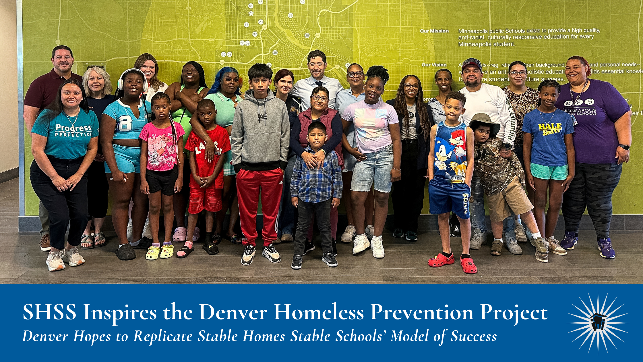 A group of adults and children pose together indoors in front of a green wall with school and city maps.