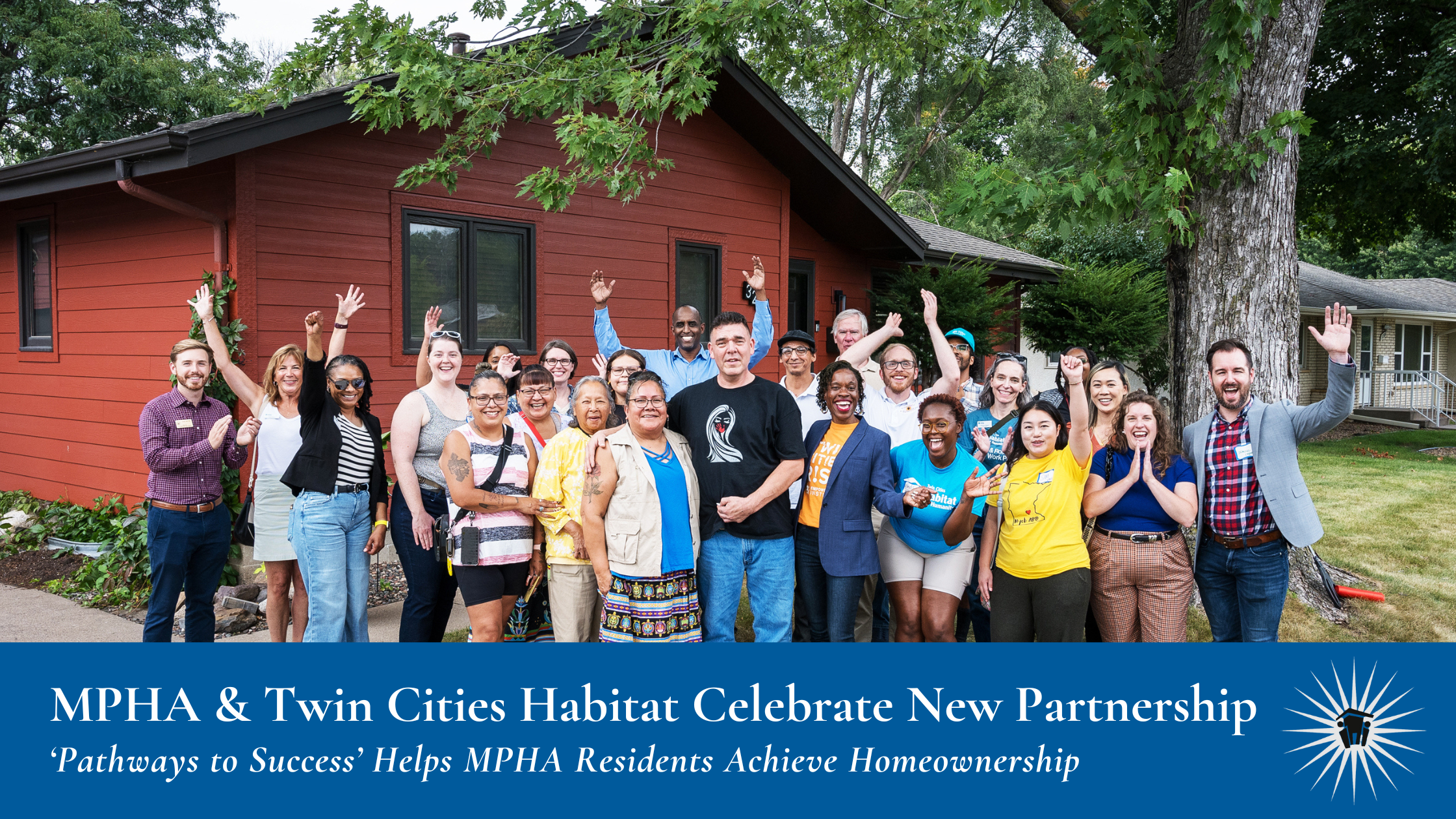 A group of adults poses and smiles for a photo outside a red house on a grassy lawn, with several people raising their hands in celebration.