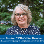 A woman with short blonde hair and glasses, wearing a navy blue patterned blouse, stands outdoors in front of green pine tree branches.