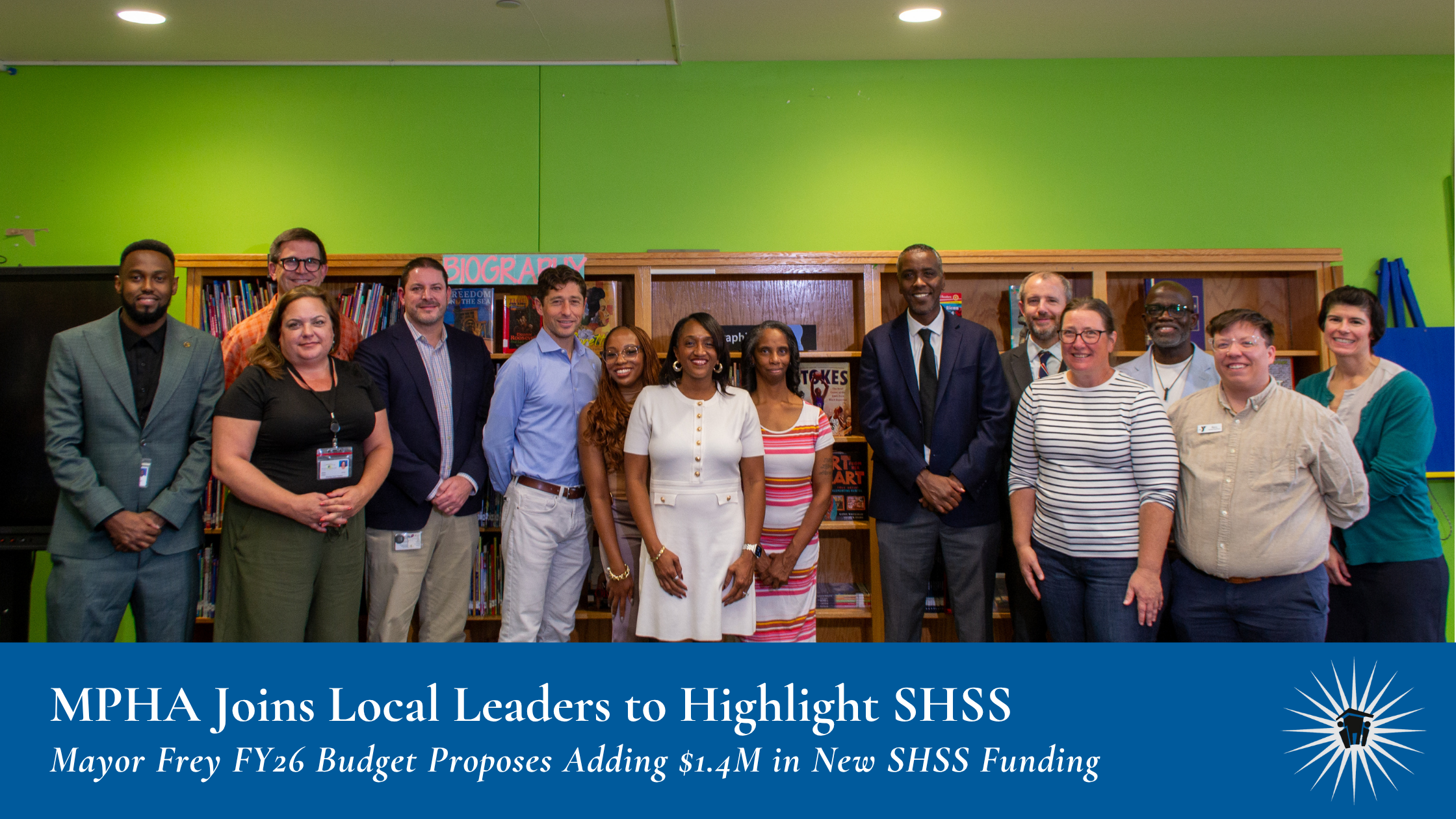 A group of adults stand together in front of bookshelves in a brightly-lit school library with green walls, posing for a group photo.