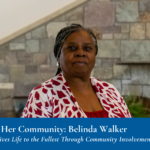 A woman stands indoors in front of a stone wall, wearing a red and white top. A blue banner at the bottom reads, "Encouraging Her Community: Belinda Walker"