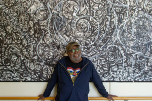 A man wearing glasses and a hat stands in front of a black-and-white abstract mural.