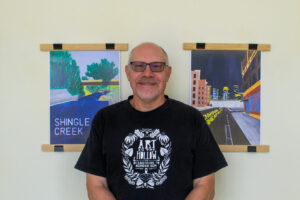 A man wearing glasses and a black t-shirt stands in front of three framed artworks on a light-colored wall.