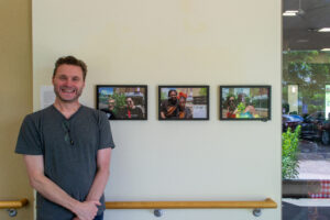 A man smiling in a gray shirt stands next to a wall with three framed photos.