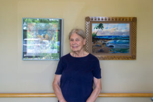 A woman, smiling softly, stands between two framed paintings on a beige wall. One painting depicts a vibrant ocean scene.