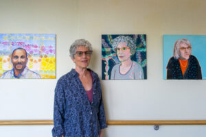 A woman stands smiling in front of three colorful portrait paintings, including one of themselves, on a light-colored wall.