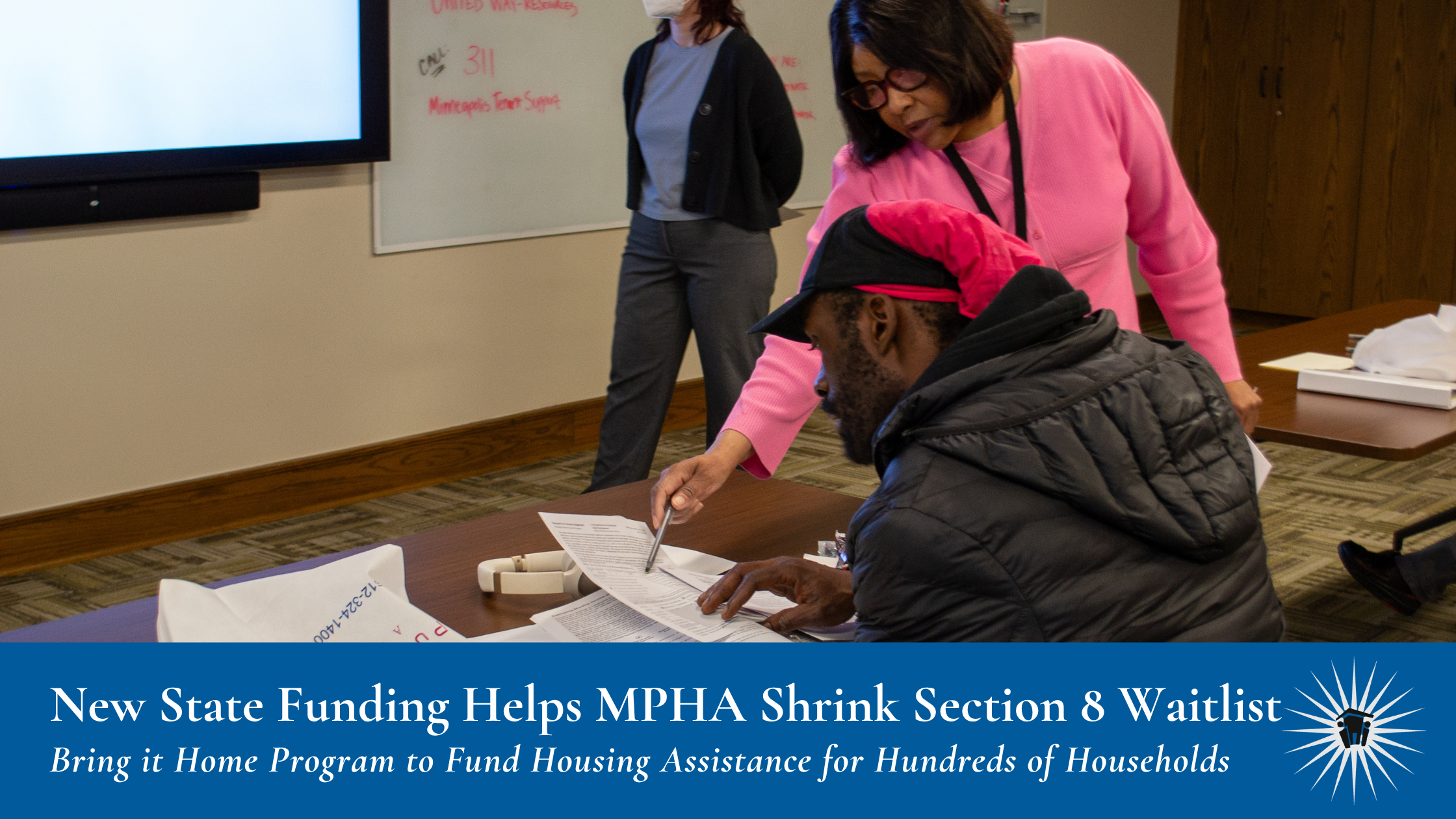 New State Funding Helps MPHA Shrink Section 8 Waitlist (1) A woman points at documents in front of a man at a table in an office setting. A text banner below reads "New State Funding Helps MPHA Shrink Section 8 Waitlist, Bring it Home Program to Fund Housing Assistance for Hundreds of Households"