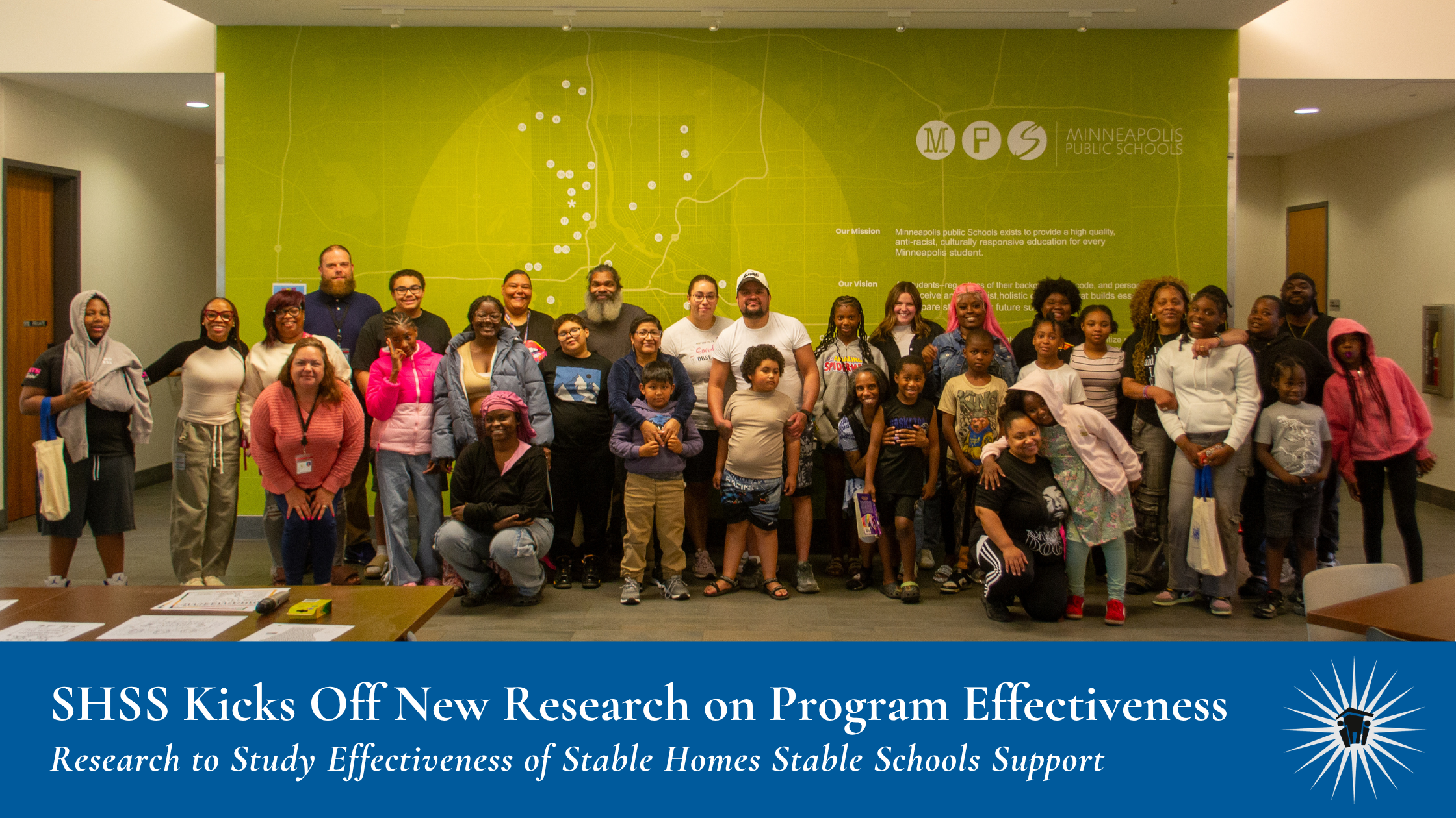 SHSS Kicks Off New Research on Program Effectiveness A couple dozen children and adults stand inside together, smiling, in front of a green wall with map. Banner at bottom reads: SHSS Kicks Off New Research on Program Effectiveness, Research to Study Effectiveness of Stable Homes Stable Schools Support