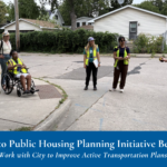 Several individuals in yellow safety vests stand talking in neighborhood walking intersection. Text banner at bottom reads: "Safe Routes to Public Housing Planning Initiative Begins, MPHA Residents Work with City to Improve Active Transportation Plans